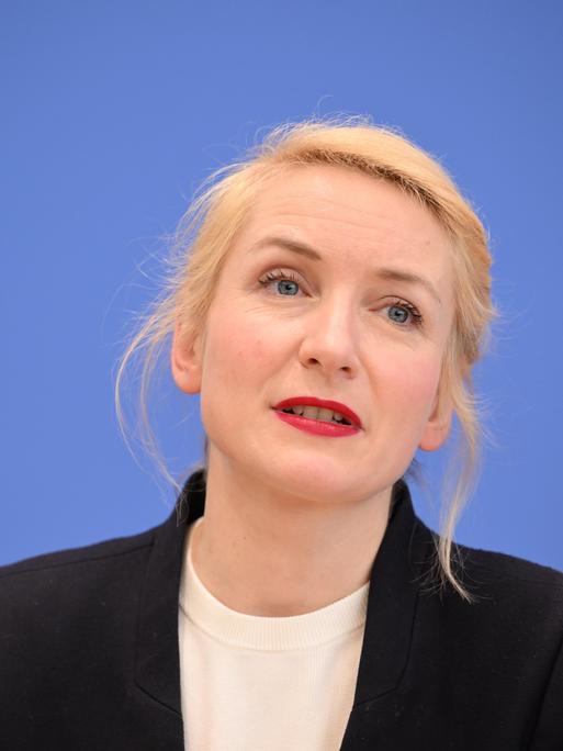 Ines Schwerdtner bei einer Pressekonferenz. Sie sitzt vor einem blauen Hintergrund und hat einen schwarzen Blazer an. 