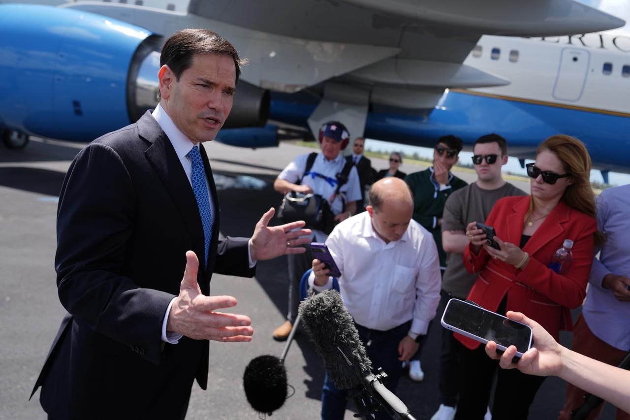 US-Außenminister Rubio steht vor Pressevertretern und spricht in Mikrofone. Er ist auf einem Flughafen. Im Hintergrund ist ein Flugzeug zu sehen. 