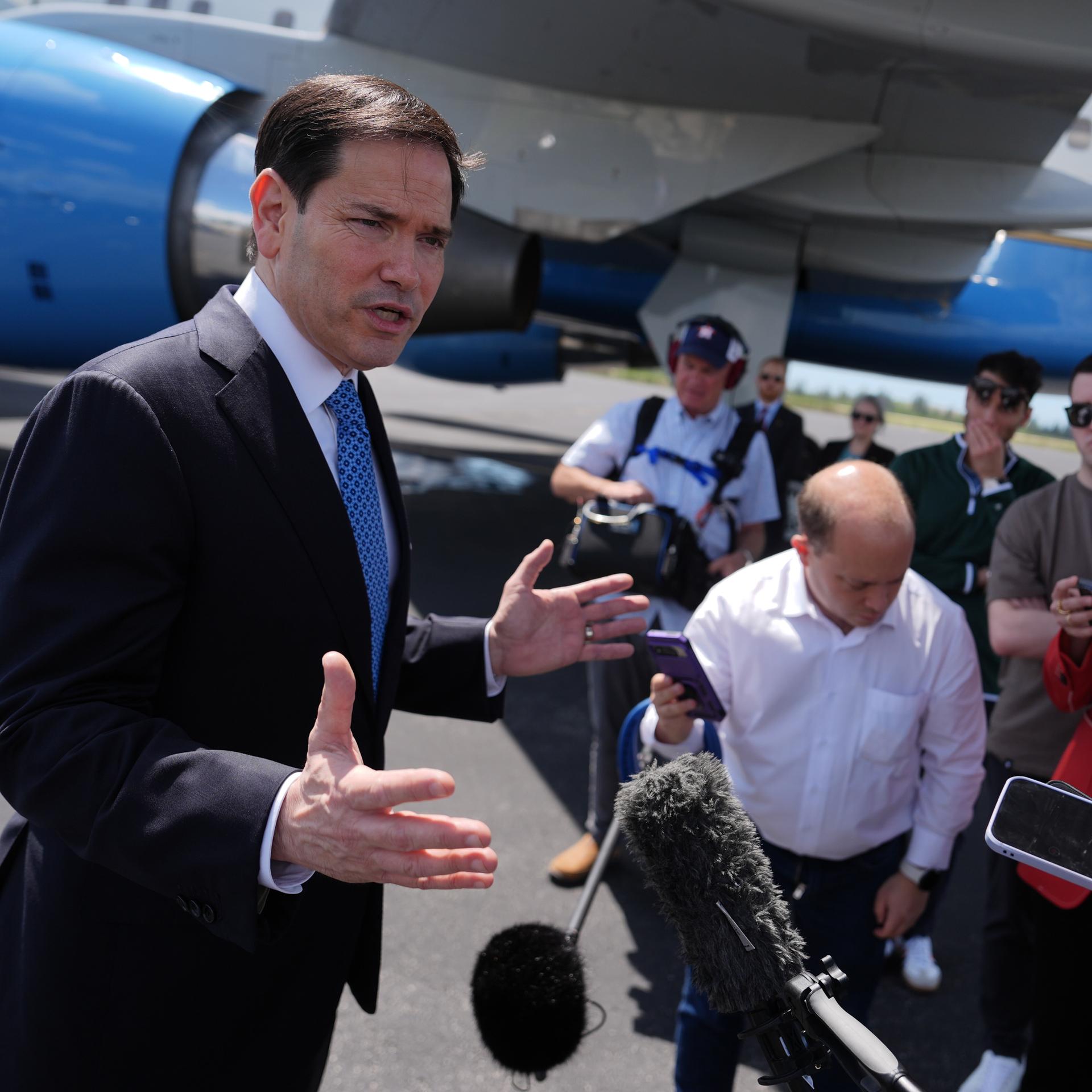 US-Außenminister Rubio steht vor Pressevertretern und spricht in Mikrofone. Er ist auf einem Flughafen. Im Hintergrund ist ein Flugzeug zu sehen. 