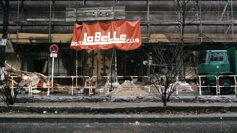 Vor dem Gebäude, in dessen Erdgeschoss sich das „La Belle“ befindet, steht ein Absperrzaun, rechts im Bild steht ein Lkw der Polizei Berlin. Vor dem Gebäude liegen Berge von Schutt. Das Gebäude ist eingerüstet, am Gerüst hängt ein Banner mit der Aufschrift „Disco – La Belle – Club“.