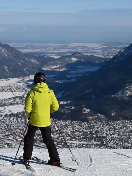 Ein Skifahrer mit Helm und gelber Jacke blickt von der Piste ins Loisachtal und auf die Häuser von Garmisch-Partenkirchen.