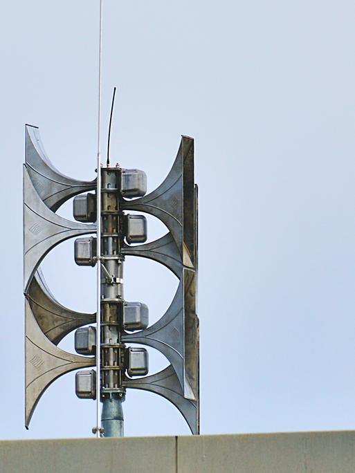 Auf einem Dach ist eine Sirenenanlage. Sie besteht aus mehreren Sirenen aus Metall, die in zwei Richtungen zeigen.