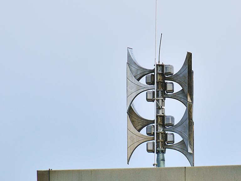 Auf einem Dach ist eine Sirenenanlage. Sie besteht aus mehreren Sirenen aus Metall, die in zwei Richtungen zeigen.