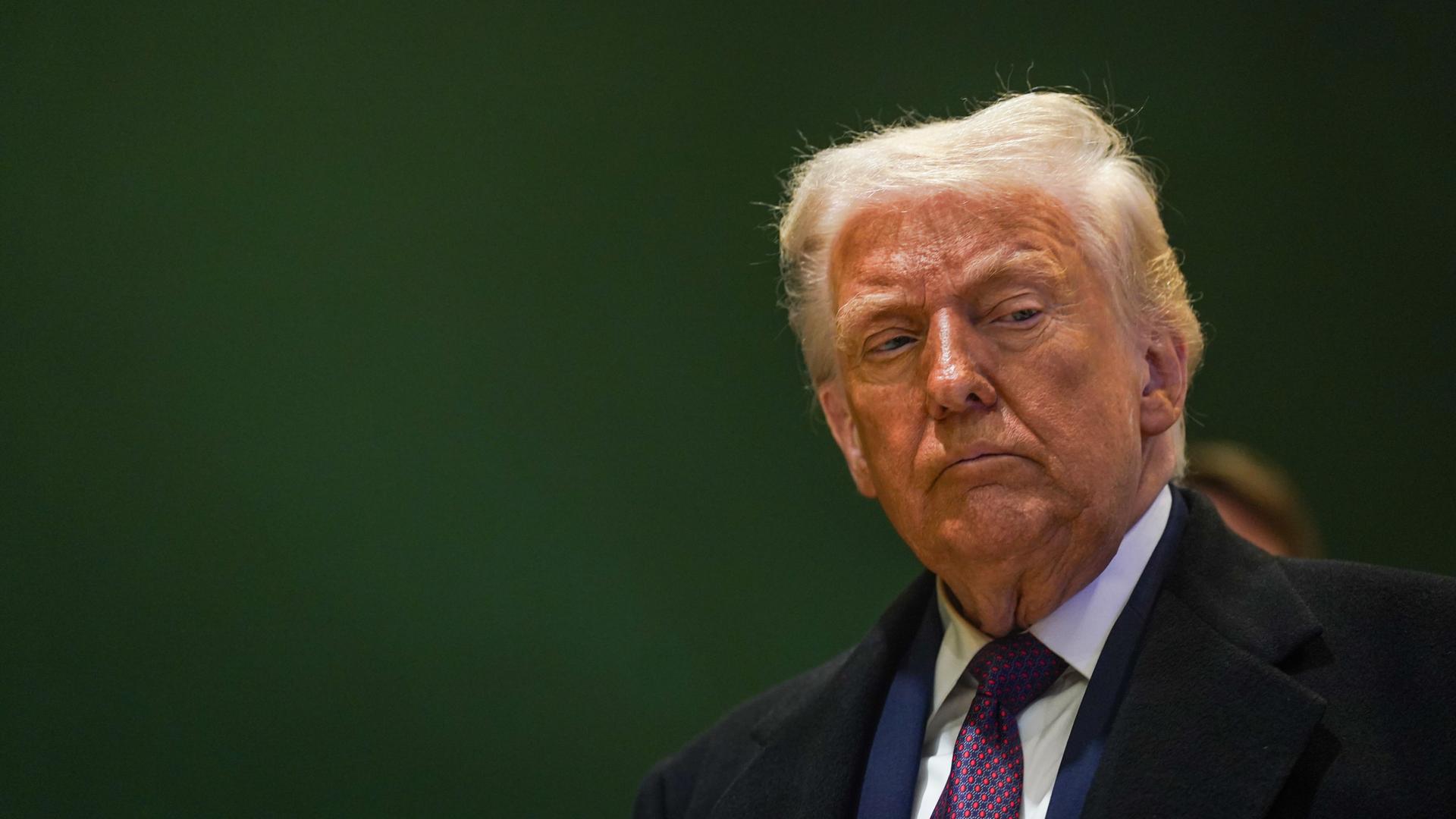 Donald Trump steht vor einer dunkelgrünen Wand und schaut nachträglich zur Seite. Donald Trump steht vor einer dunkelgrünen Wand und schaut nachträglich zur Seite.