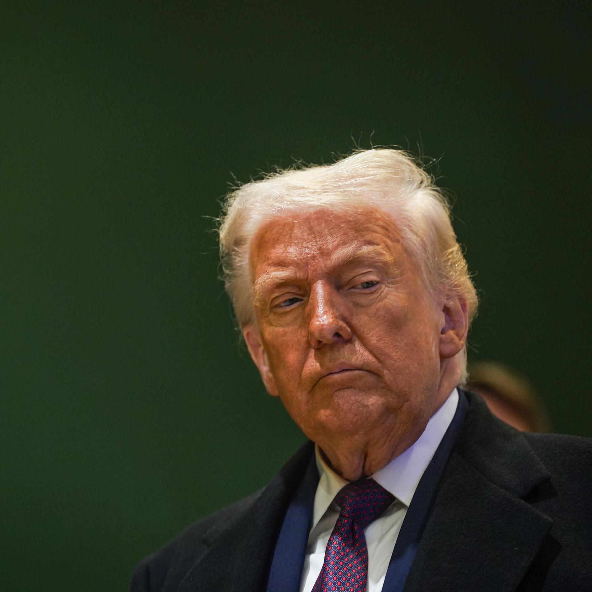 Donald Trump steht vor einer dunkelgrünen Wand und schaut nachträglich zur Seite.