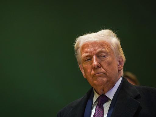 Donald Trump steht vor einer dunkelgrünen Wand und schaut nachträglich zur Seite.