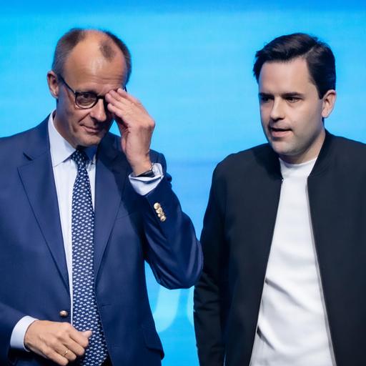 Bundeskanzler Friedrich Merz steht neben Johannes Winkel, Bundesvorsitzender Jungen Union, und fasst sich an die Brille, beide sehen nicht sehr glücklich aus