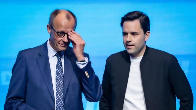 Bundeskanzler Friedrich Merz steht neben Johannes Winkel und fasst sich an die Brille.