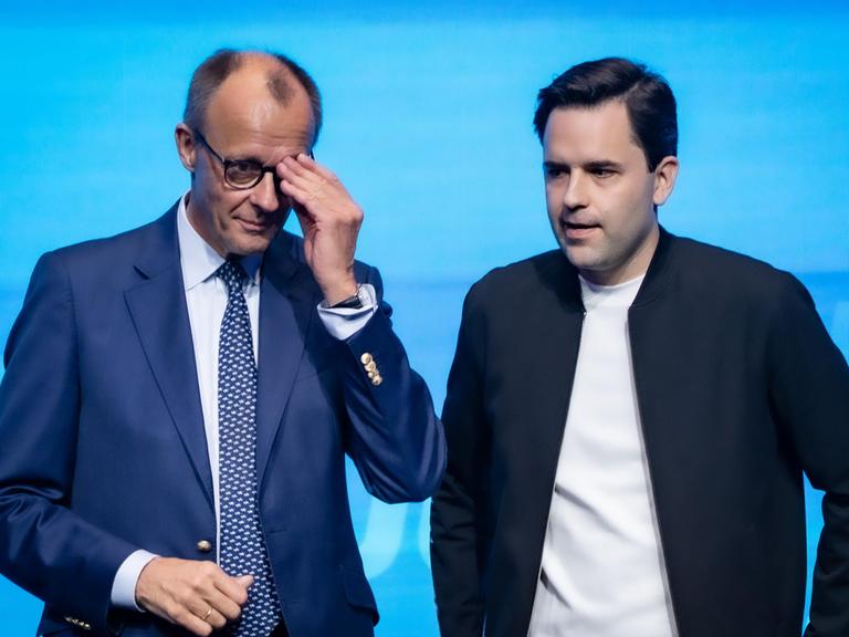 Bundeskanzler Friedrich Merz steht neben Johannes Winkel, Bundesvorsitzender Jungen Union, und fasst sich an die Brille, beide sehen nicht sehr glücklich aus Bundeskanzler Friedrich Merz steht neben Johannes Winkel, Bundesvorsitzender Jungen Union, und fasst sich an die Brille, beide sehen nicht sehr glücklich aus