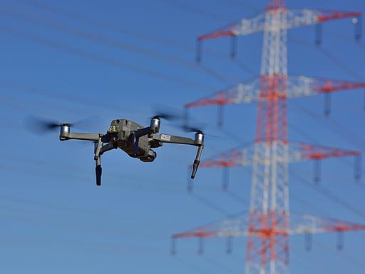 Eine Drohne fliegt in der Luft, dahinter sind Stommasten zu sehen.