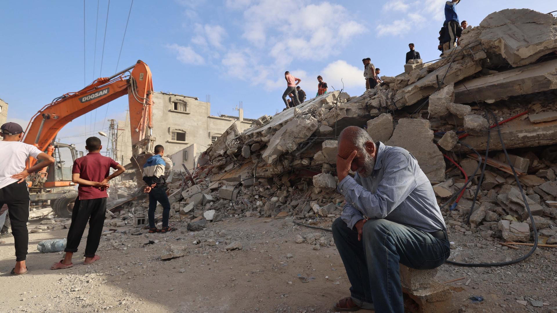Zu sehen ist eine Szene in der Stadt Rafah im Gaza-Streifen. Ein Bagger räumt Trümmer nach einem Angriff weg. Ein Mann sitzt im Vordergrund und trauert.