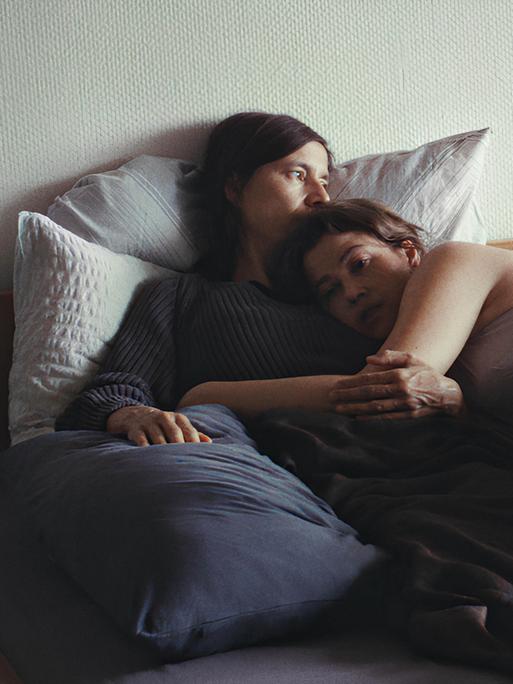 Zwei Frauen liegen im Bett. Die eine ist angezogen und hält eine traurig wirkende, nur in Unterwäsche bekleidete Frau im Arm.