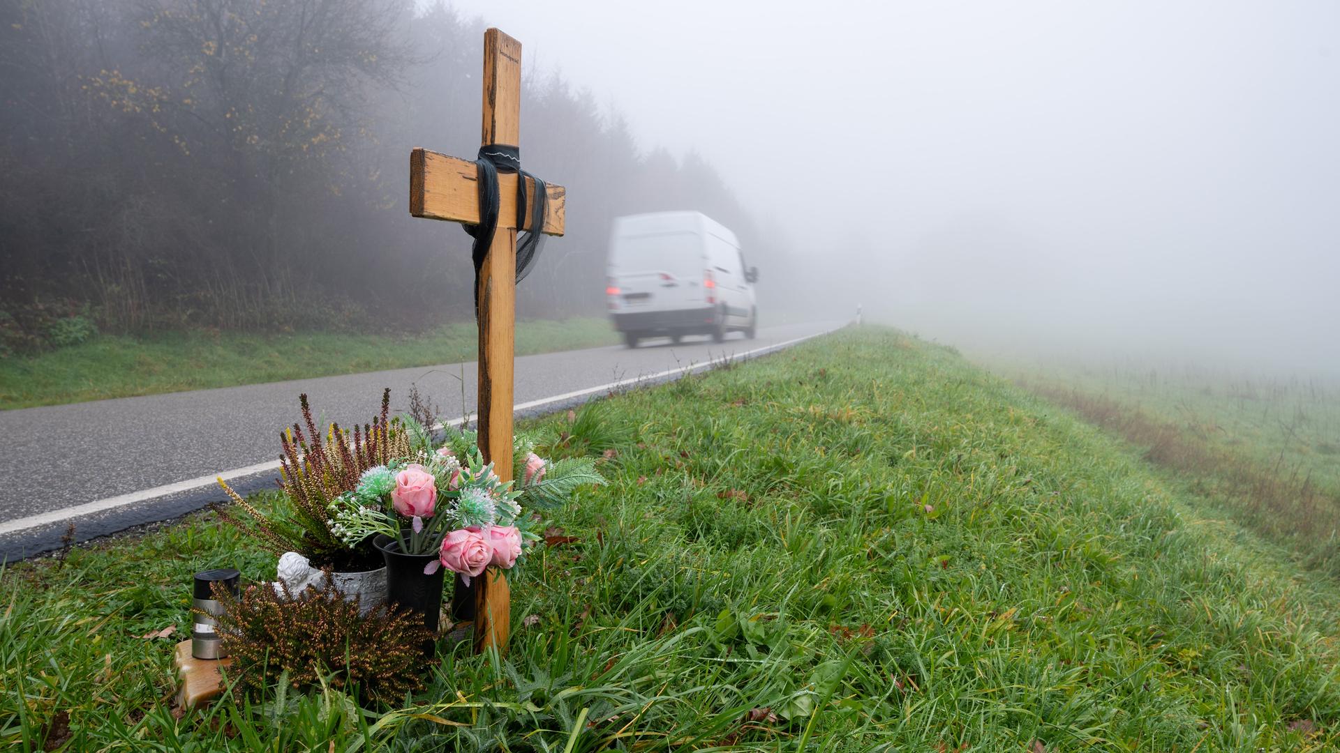 Das Holzkreuz mit Blumen steht am Straßenrand im Bildvordergrund. Dahinter ein weißer Transporter, der auf der Straße in eine Nebelwand fährt. Das Holzkreuz mit Blumen steht am Straßenrand im Bildvordergrund. Dahinter ein weißer Transporter, der auf der Straße in eine Nebelwand fährt.
