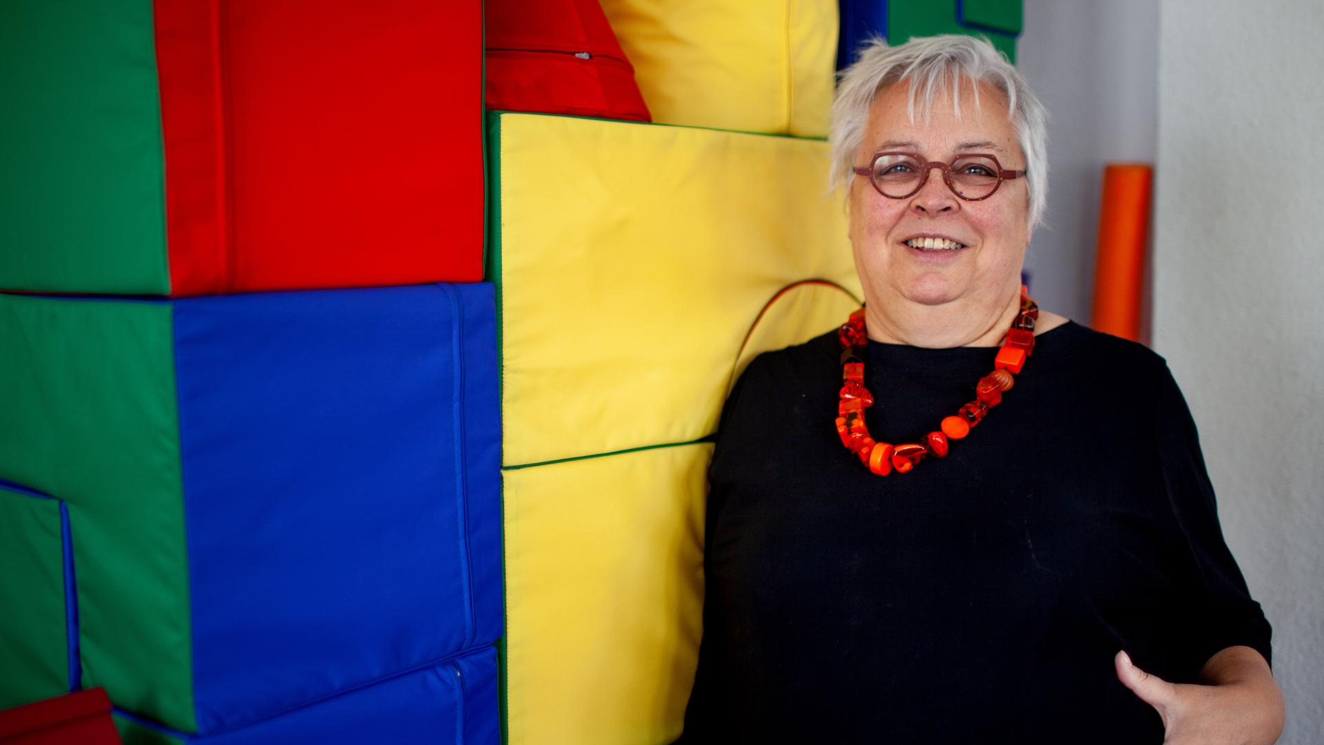 Die Erziehungswissenschaftlerin und Diplom-Pädagogin Ursula Enders trägt ein schwarzes Kleid, kurze Haare und eine Brille. Sie lehnt an einer Wand aus farbigen Stoffwürfeln und lacht gestikulierend in die Kamera.