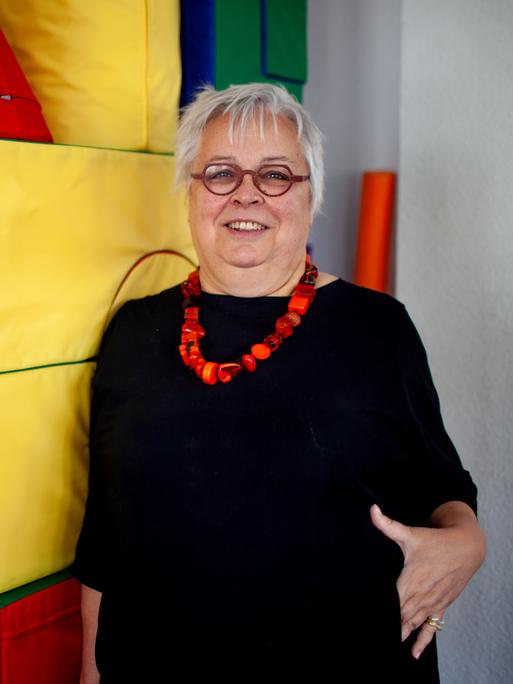 Die Erziehungswissenschaftlerin und Diplom-Pädagogin Ursula Enders trägt ein schwarzes Kleid, kurze Haare und eine Brille. Sie lehnt an einer Wand aus farbigen Stoffwürfeln und lacht gestikulierend in die Kamera.