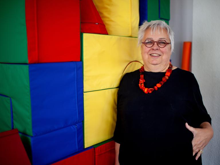 Die Erziehungswissenschaftlerin und Diplom-Pädagogin Ursula Enders trägt ein schwarzes Kleid, kurze Haare und eine Brille. Sie lehnt an einer Wand aus farbigen Stoffwürfeln und lacht gestikulierend in die Kamera.