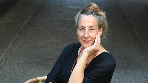 Die Schriftstellerin Judith Hermann sitzt draußen auf einem gepflasterten Platz auf einem Schaukelstuhl und stützt ihr Kinn auf eine Hand.