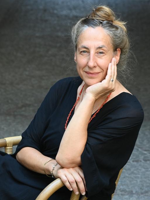 Die Schriftstellerin Judith Hermann sitzt draußen auf einem gepflasterten Platz auf einem Schaukelstuhl und stützt ihr Kinn auf eine Hand.