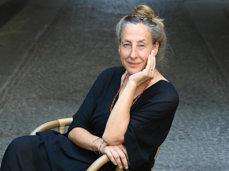 Die Schriftstellerin Judith Hermann sitzt draußen auf einem gepflasterten Platz auf einem Schaukelstuhl und stützt ihr Kinn auf eine Hand.