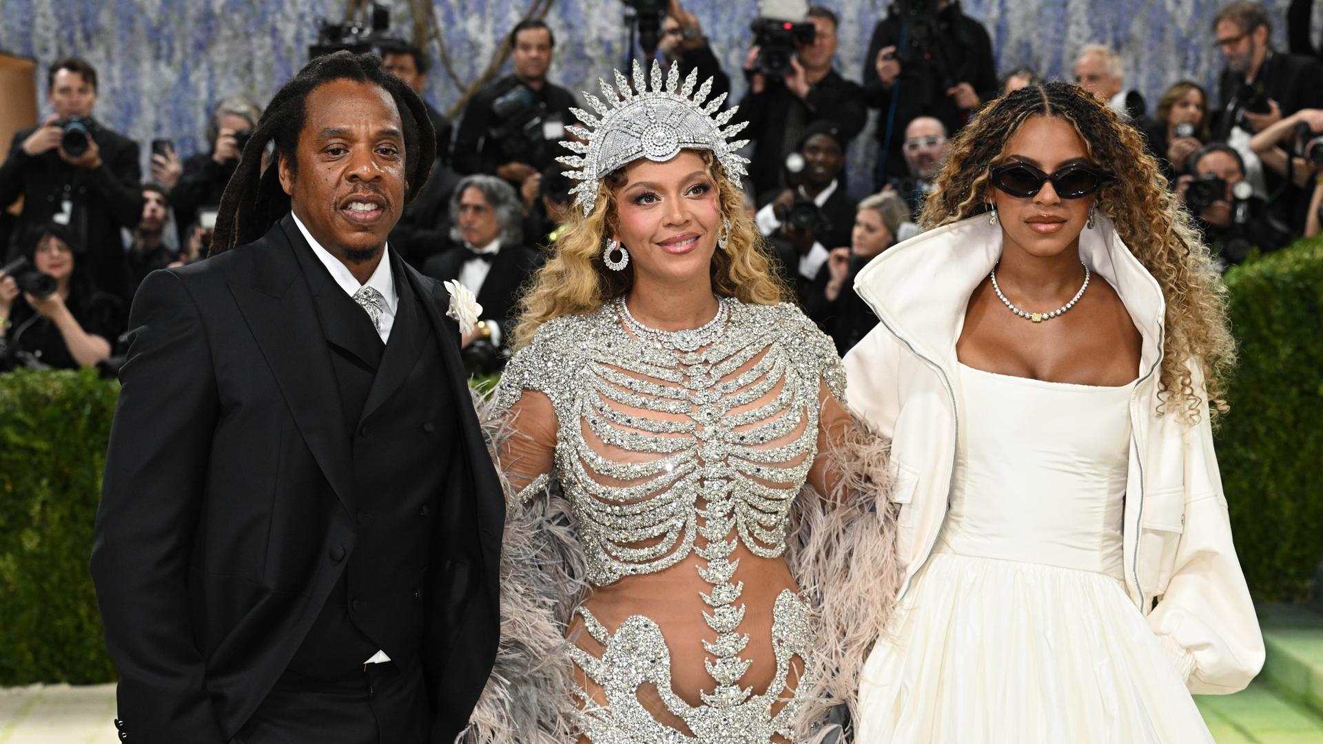 Jay-Z im schwarzen Anzug, Beyonce und Blue Ivy Carter in aufwändigen Kostümen stehen vor einer Schar Fotografen.