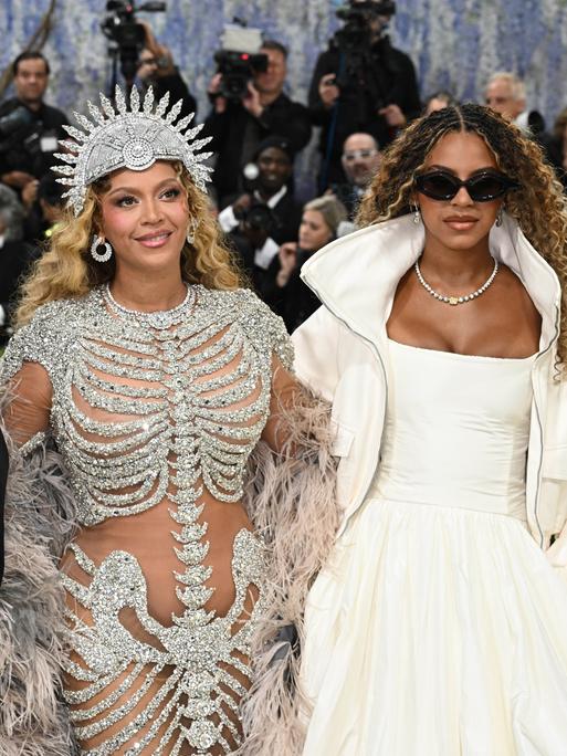 Jay-Z im schwarzen Anzug, Beyonce und Blue Ivy Carter in aufwändigen Kostümen stehen vor einer Schar Fotografen.