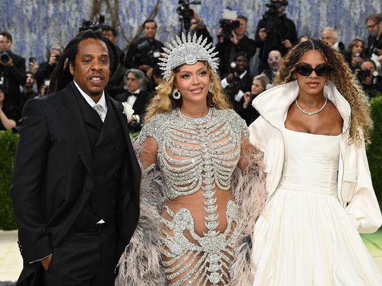 Jay-Z im schwarzen Anzug, Beyonce und Blue Ivy Carter in aufwändigen Kostümen stehen vor einer Schar Fotografen.