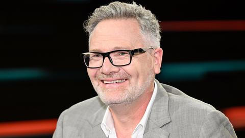 Der britische Wirtschaftshistoriker Adam Tooze sitzt in einem Fernsehstudio vor einen dunklen Hintergrund. Er hat einen grauen Vollbart und trägt einen hellgrauen Anzug.

