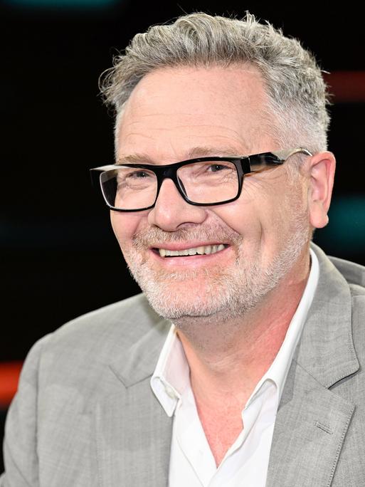 Der britische Wirtschaftshistoriker Adam Tooze sitzt in einem Fernsehstudio vor einen dunklen Hintergrund. Er hat einen grauen Vollbart und trägt einen hellgrauen Anzug.
