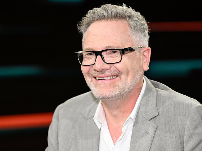 Der britische Wirtschaftshistoriker Adam Tooze sitzt in einem Fernsehstudio vor einen dunklen Hintergrund. Er hat einen grauen Vollbart und trägt einen hellgrauen Anzug.
