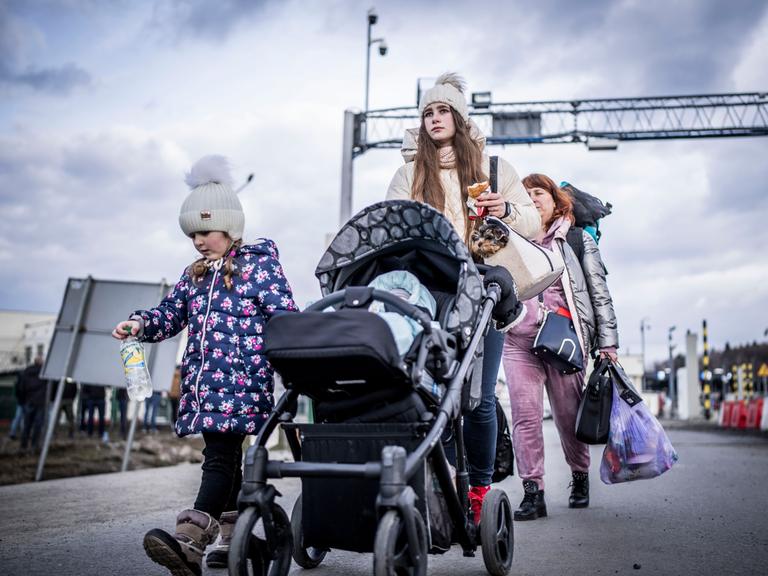 Ein Mädchen schiebt einen Kinderwagen, ein anderes läuft neben ihr her. Ihnen folgt eine Frau, die mehrere Tasche trägt. Im Hintergrund ist eine Grenzanlage zu sehen.