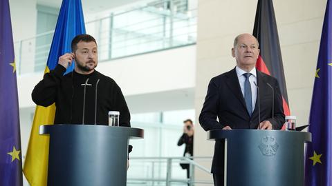 Der ukrainische Präsident Selenskyj und Bundeskanzler Scholz bei einer Pressekonferenz in Berlin. Beide stehen an Rednerpulten vor Flaggen Deutschlands, der Ukraine und der Europäischen Union.