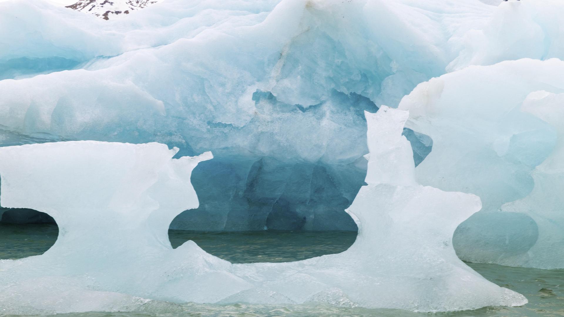 Blaues Gletschereis von einem großen Eisberg, der im Meeer schwimmt. Davor ein schon stark abgeschmolzenes kleines Eisstück.