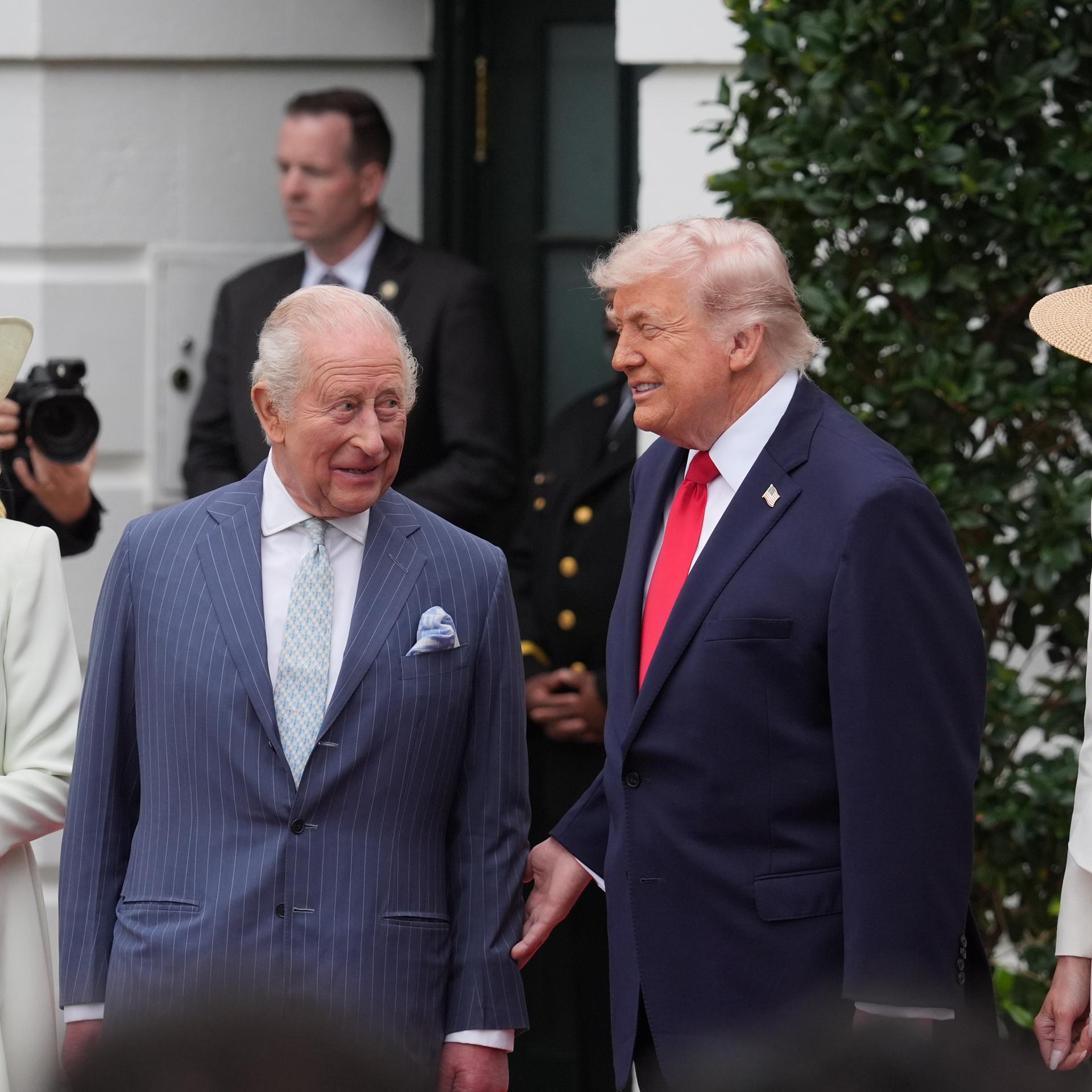 König Charles und US-Präsident Trump stehen vor dem Weißen Haus und reden miteinander. Neben Charles steht Camilla, in einem weißen Kleid und mit einem großen weißen Hut.