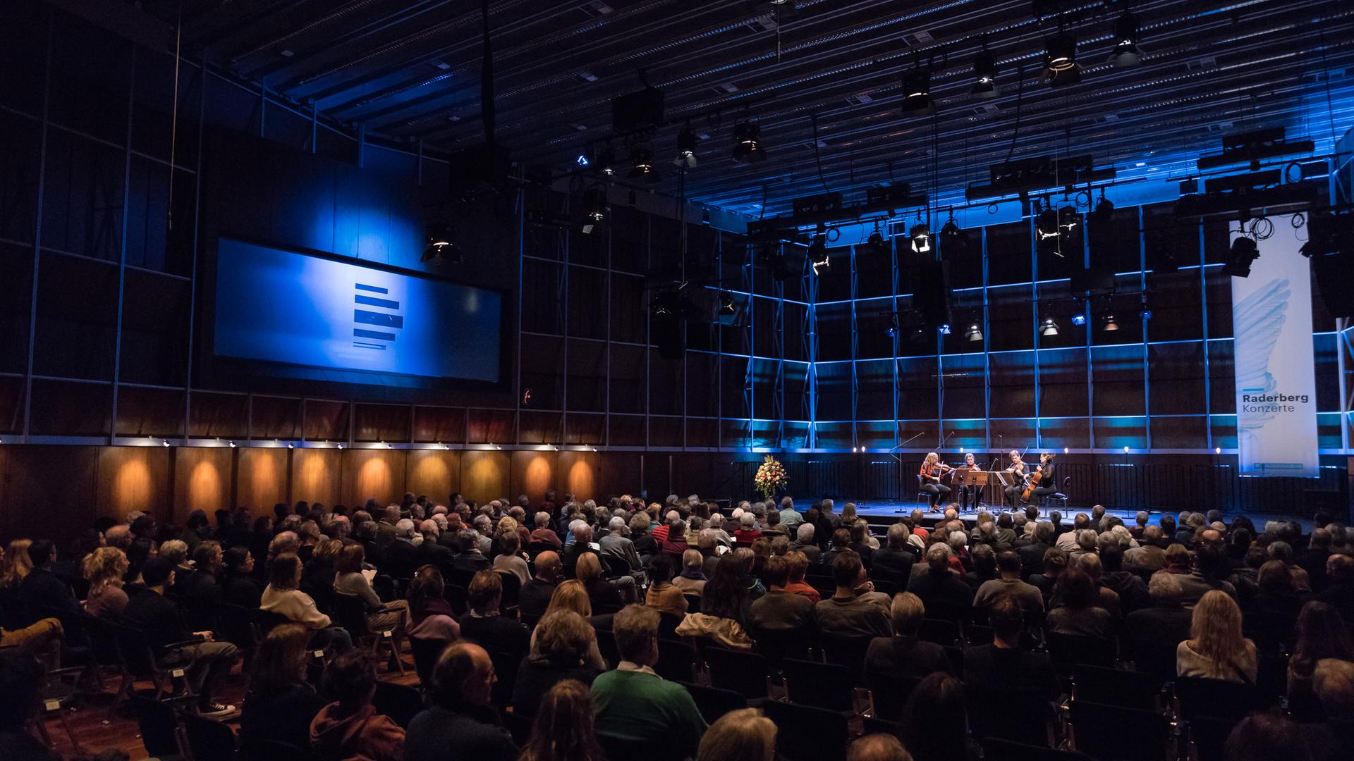 Totale eines bläulich erleuchteten Konzertsaals mit voll besetzten Stuhlreihen. Vorne auf der Bühne sitzt rechter Hand ein Streichquartett. Direkt daneben steht ein Banner des Deutschlandfunk mit der Aufschrift "Raderbergkonzert". Links im Saal ist das Logo des Dlf an der Wand zu sehen.