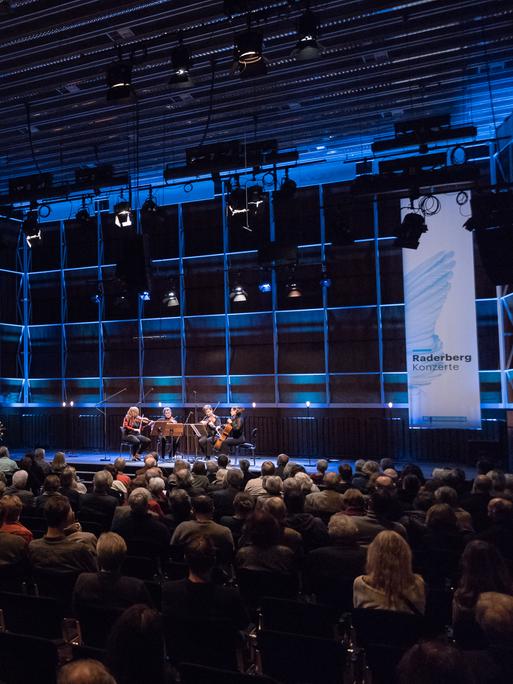 Totale eines bläulich erleuchteten Konzertsaals mit voll besetzten Stuhlreihen. Vorne auf der Bühne sitzt rechter Hand ein Streichquartett. Direkt daneben steht ein Banner des Deutschlandfunk mit der Aufschrift "Raderbergkonzert". Links im Saal ist das Logo des Dlf an der Wand zu sehen.