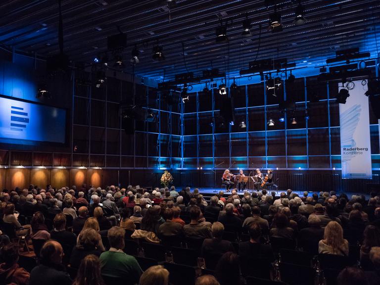 Totale eines bläulich erleuchteten Konzertsaals mit voll besetzten Stuhlreihen. Vorne auf der Bühne sitzt rechter Hand ein Streichquartett. Direkt daneben steht ein Banner des Deutschlandfunk mit der Aufschrift "Raderbergkonzert". Links im Saal ist das Logo des Dlf an der Wand zu sehen.