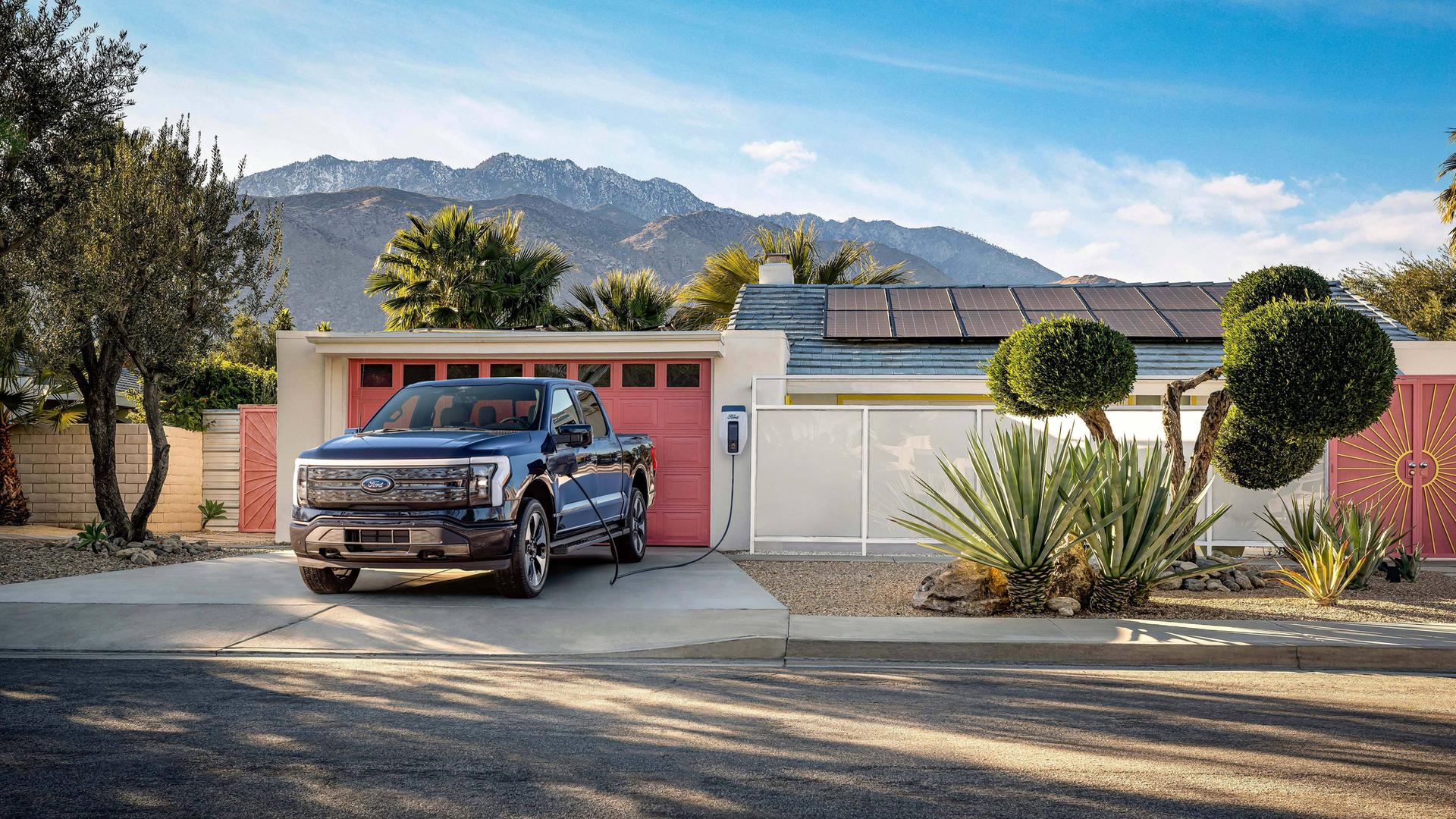 Ein Ford F-150 Lightning Pickup-Truck steht vor einem typischen amerikanischen Haus mit Garage, wo er aufgeladen wird. Im Hintergrund sind berge und Palem zu sehen.