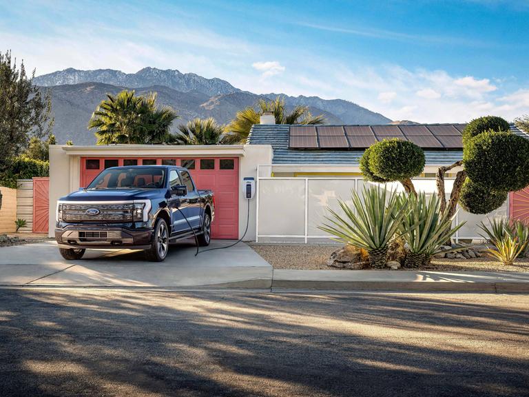 Ein Ford F-150 Lightning Pickup-Truck steht vor einem typischen amerikanischen Haus mit Garage, wo er aufgeladen wird. Im Hintergrund sind berge und Palem zu sehen.
