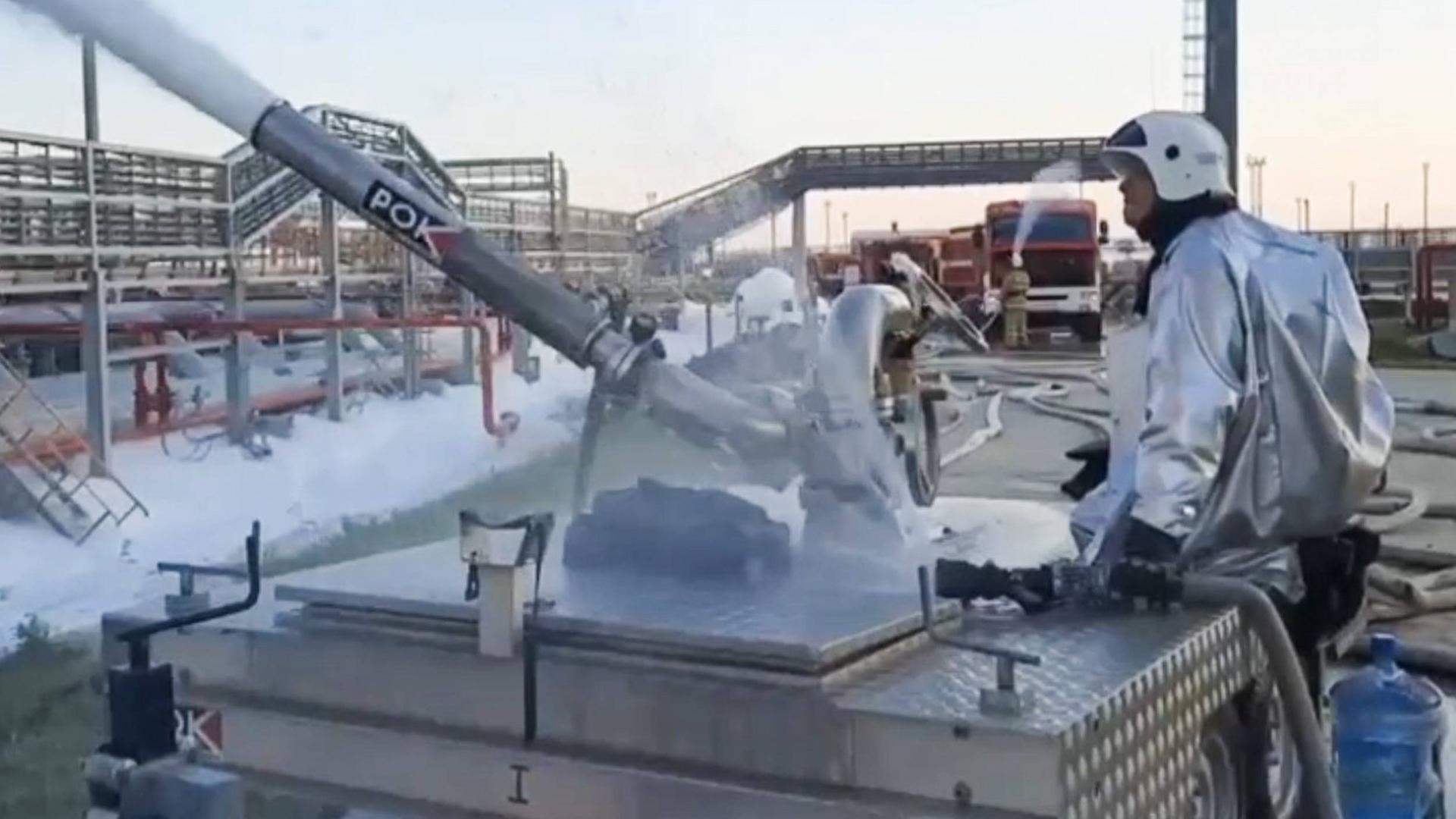 Ein Feuerwehrmann an einem Löschgerät in einem Öldepot in Russland. Im Hintergrund Feuerwagen und Rauch, der aus den Leitungen steigt.