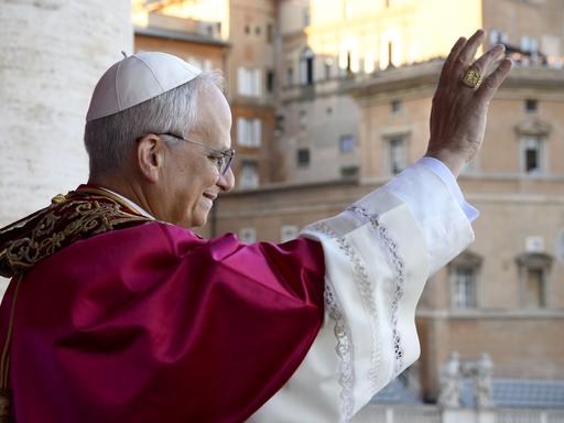 Leo XIV winkt lächelnd vom Balkon des Petersdoms. Er ist von der Seite fotografiert.