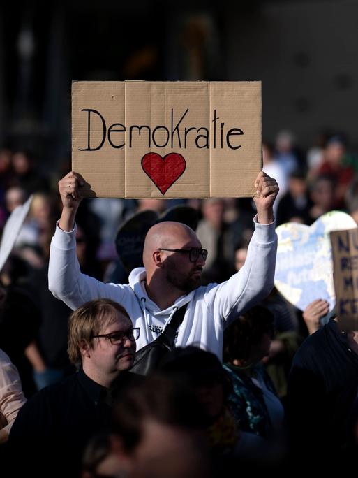 Ein Man hält auf einer Demo ein Schild in die Höhe, auf dem "Demokratie" steht. Darunter ist ein rotes Herz gemalt.