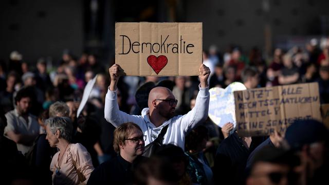 Ein Man hält auf einer Demo ein Schild in die Höhe, auf dem "Demokratie" steht. Darunter ist ein rotes Herz gemalt.