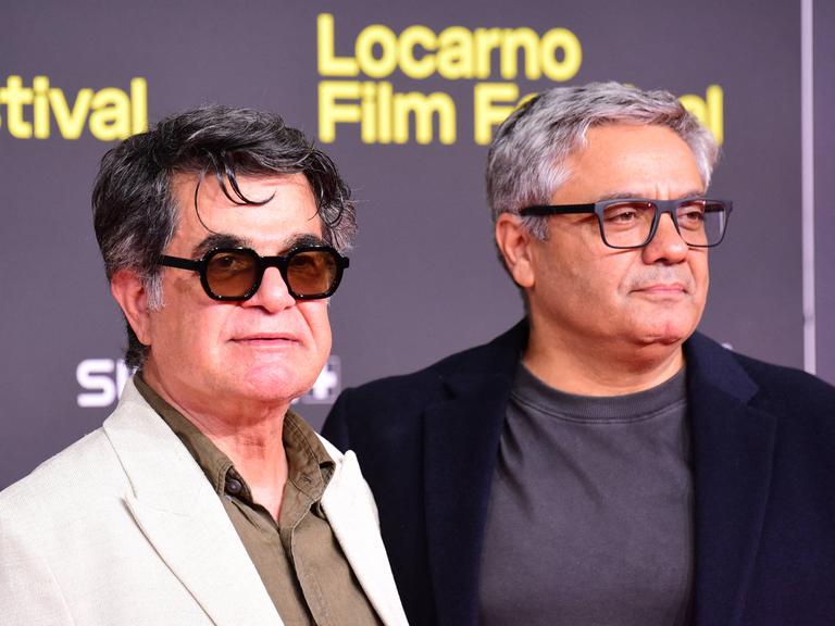 Jafar Panahi und Mohammad Rasoulof stehen nebeneinander vor einer Wand auf der "Locarno Filmfestival" steht. 