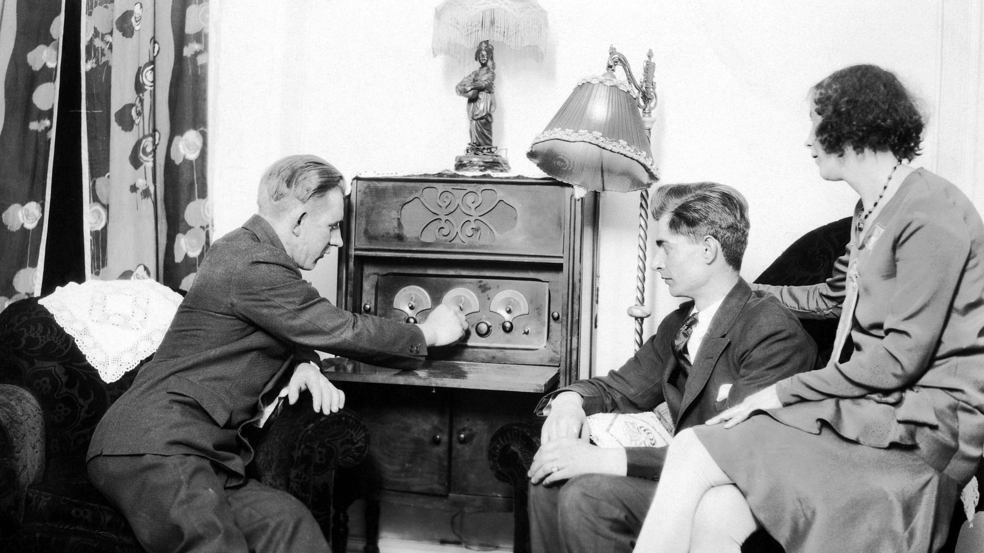 Zwei Männer und eine Frau sitzen in den 1920er Jahren in einem Wohnzimmer vor einem Radiogerät, dass einer der Männer gerade bedient. 