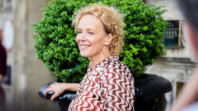 Das Bild zeigt die Schauspielerin Katja Riemann. Sie steht seitlich zur Kamera, hinter ihrem Kopf ist ein grüner Buchsbaum zu sehen. Sie trägt ein Kleid, auf dem sich dunkelrote Linien über weißen Grund schlängeln. 