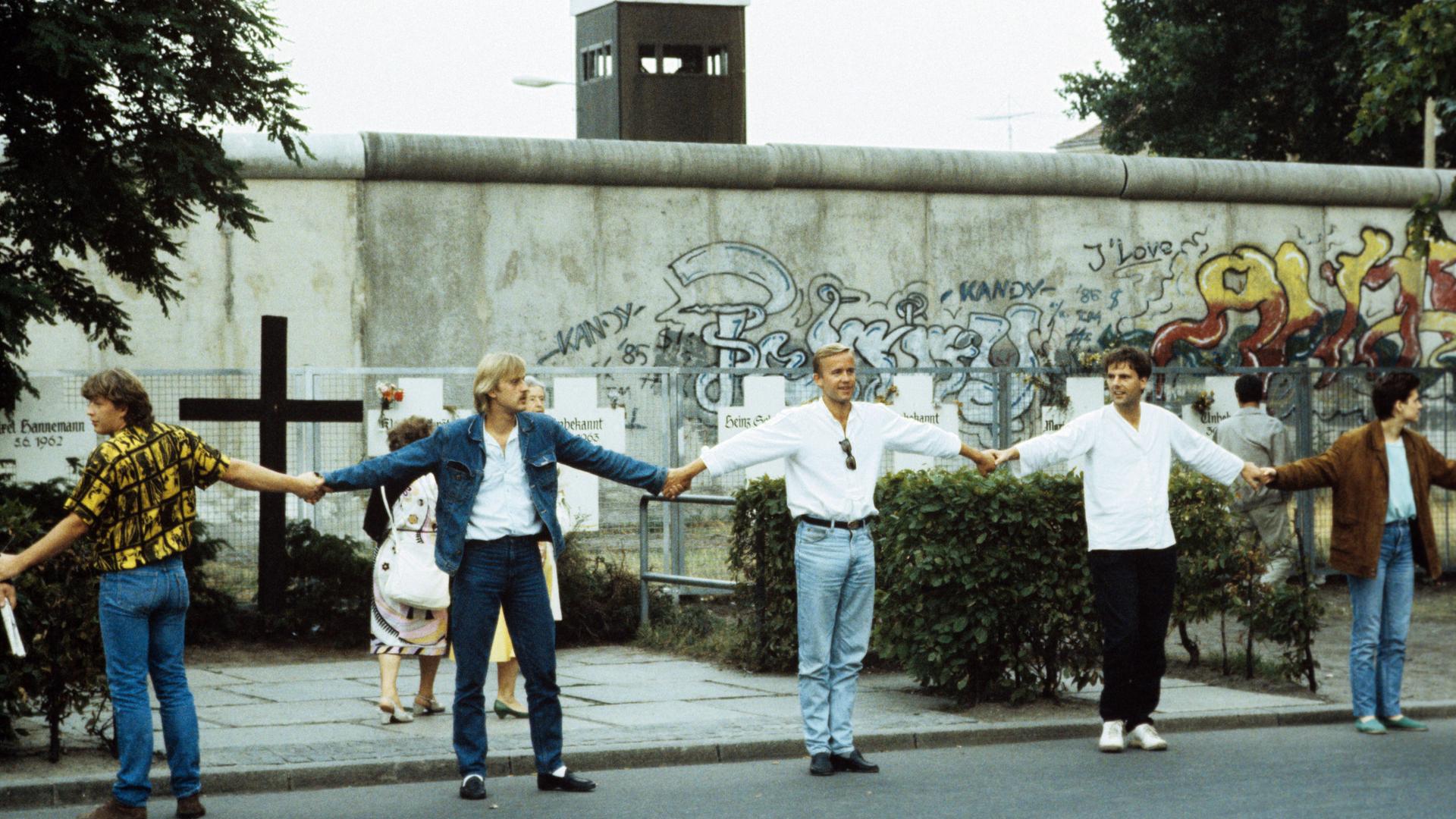 Eine Menschenkette an der Westberliner Mauer. Im Hintergrund ist ein Gedenkkreuz für die Mauertoten zu sehen.  Eine Menschenkette an der Westberliner Mauer. Im Hintergrund ist ein Gedenkkreuz für die Mauertoten zu sehen.