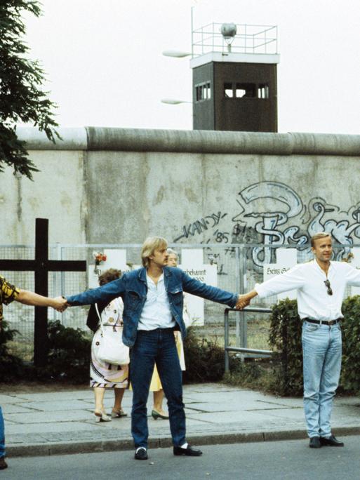 Eine Menschenkette an der Westberliner Mauer. Im Hintergrund ist ein Gedenkkreuz für die Mauertoten zu sehen.  Eine Menschenkette an der Westberliner Mauer. Im Hintergrund ist ein Gedenkkreuz für die Mauertoten zu sehen.