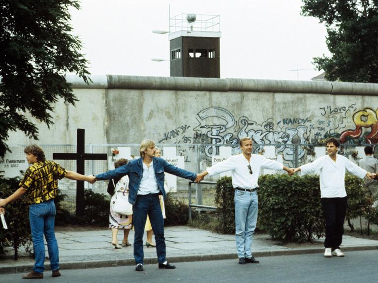 Eine Menschenkette an der Westberliner Mauer. Im Hintergrund ist ein Gedenkkreuz für die Mauertoten zu sehen.  Eine Menschenkette an der Westberliner Mauer. Im Hintergrund ist ein Gedenkkreuz für die Mauertoten zu sehen.