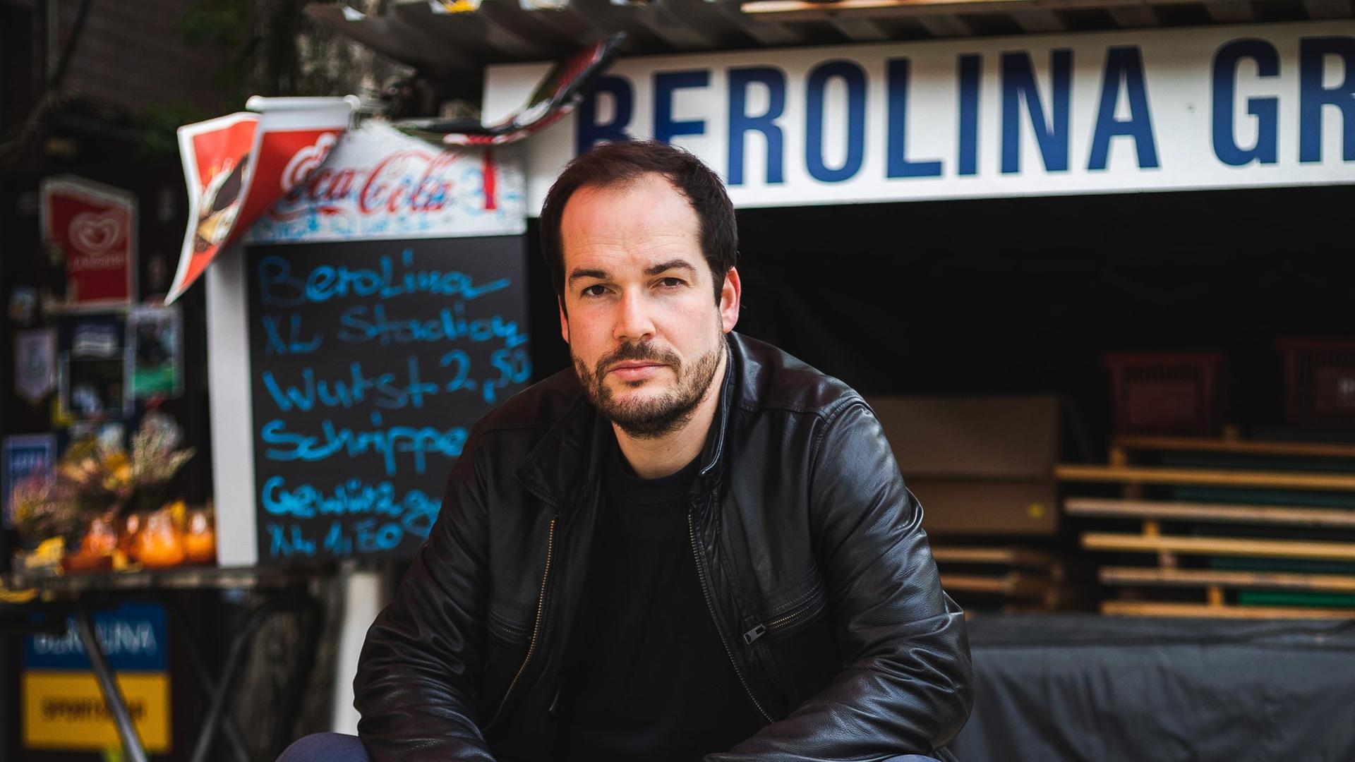Ein Mann mit kurzen, dunklen Haaren sitzt in dunkler Lederjacke auf dem Tisch einer Bierbank in einem geschlossenen Berliner Biergarten. Ein Mann mit kurzen, dunklen Haaren sitzt in dunkler Lederjacke auf dem Tisch einer Bierbank in einem geschlossenen Berliner Biergarten.