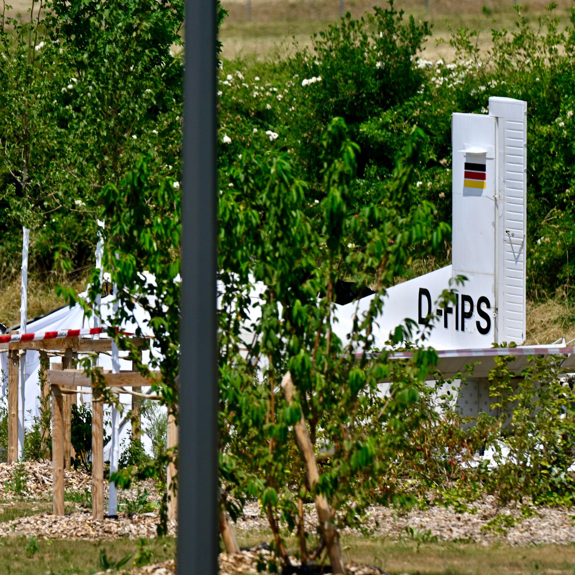 Zwischen Bäumen liegt der zerstörte Rumpf des weißen Flugzeugs. Er ist teilweise mit einer Plane abgedeckt.